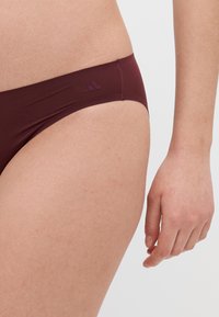 Chiloție bikini burgund cu textură netedă, design cu talie joasă și detaliu logo subtil la brâu. Materialul pare elastic și se potrivește strâns.