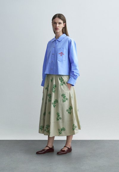 Γυναίκα που φοράει ανοιχτό μπλε πουκάμισο με κουμπιά και λογότυπο στην τσέπη, μπεζ φούστα με πράσινο λουλουδάτο σχέδιο και καφέ παπούτσια Mary Jane, στέκεται μέσα σε εσωτερικό χώρο.