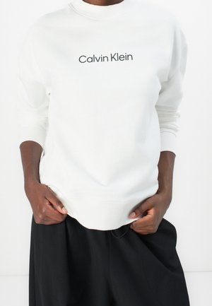 Personne à la peau foncée portant un sweat blanc Calvin Klein et un pantalon noir, les mains saisissant les bords inférieurs du sweat.