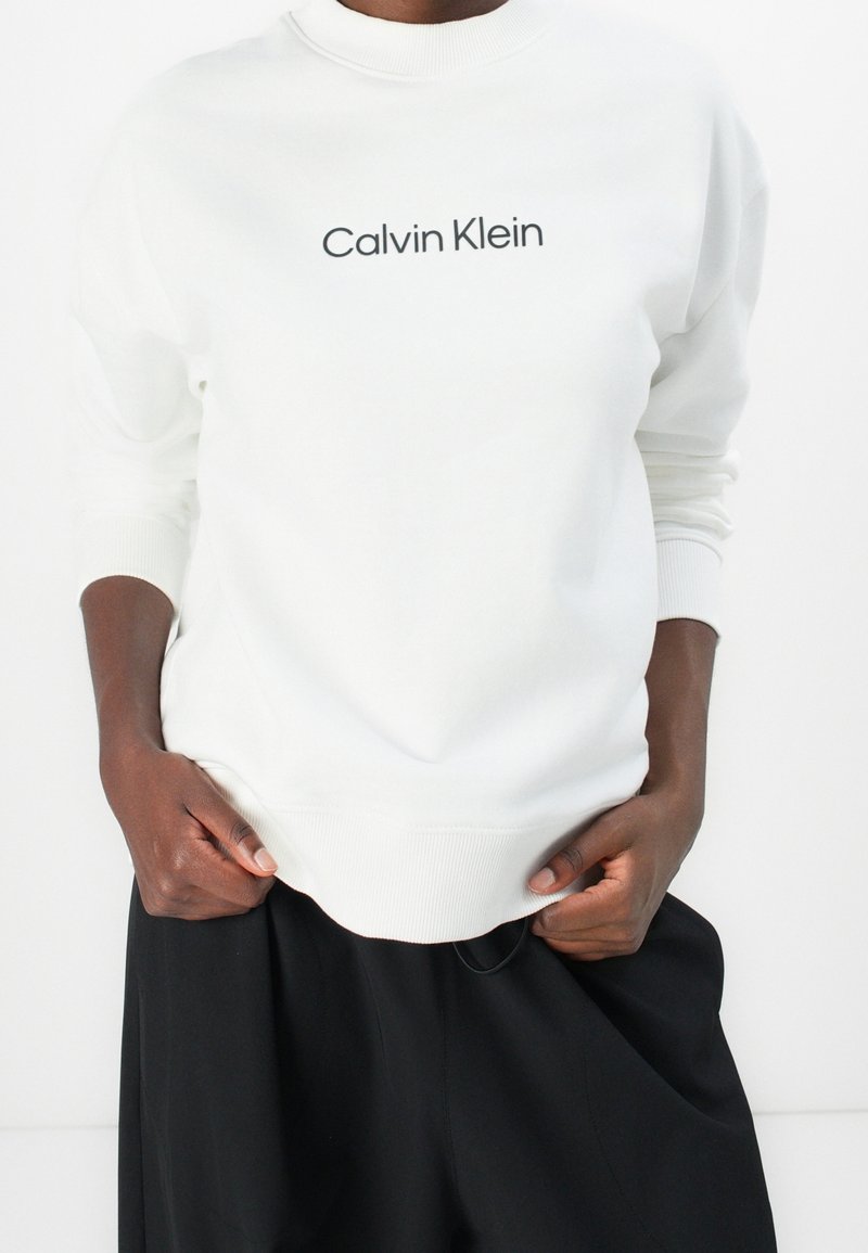 Personne à la peau foncée portant un sweat blanc Calvin Klein et un pantalon noir, les mains saisissant les bords inférieurs du sweat.