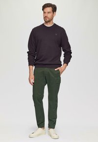 Man in een zwart sweatshirt met lange mouwen, groene cargobroek met zijzakken en witte sneakers, staand met één hand in de zak.