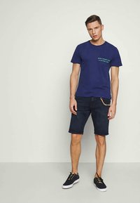 Navy blue cotton t-shirt with green text on the chest, paired with dark denim shorts and black sneakers. Simple design and casual fit.