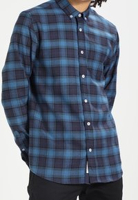 Koszula w kratę w odcieniach niebieskiego i granatowego, z bawełnianego materiału, z kołnierzykiem, guzikami z przodu i kieszonką. Tkanina ma gładką, miękką fakturę.
