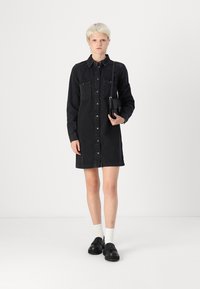Black denim shirt dress with a button-down front and two chest pockets. Worn with black shoes and white ankle socks. Simple, tailored design.
