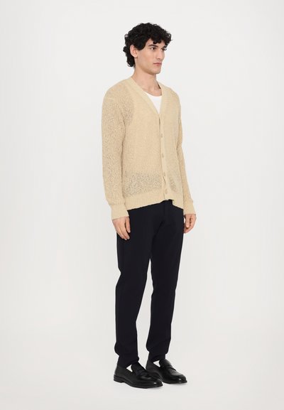 Beige strikket cardigan med V-hals og frontknapper, brukt over en hvit skjorte, kombinert med marineblå bukser og svarte slip-on sko.