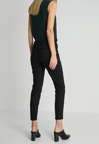 Pantalons noirs ajustés avec une texture lisse, un design fuselé et des poches latérales. Associés à un haut sans manches foncé et des mules à talons noirs.