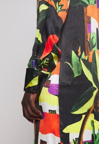 Main à la peau foncée reposant à côté d’un vêtement à manches longues coloré avec des motifs audacieux de feuilles et abstraits en vert, orange, violet et noir.