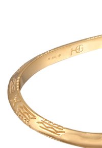 Guldpläterat metallarmband med strukturerad yta som har abstrakta mönster, graverad logotyp och stämplade detaljer. Rund form.