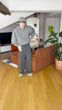 Šedá mikina na zip, šedé kalhoty na míru a stříbrné tenisky. Stojí v místnosti s dřevěnou podlahou a texturovanou oranžovou pohovkou.