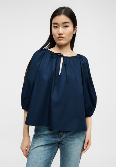 Νεαρή γυναίκα με μακριά σκούρα μαλλιά, φοράει χαλαρό μπλε πουκάμισο με μανίκια τριών τετάρτων και ανοιχτόχρωμα τζιν, στέκεται μπροστά σε ένα απλό φόντο.