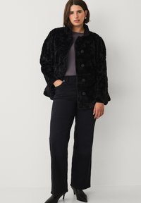 Sort faux fur frakke med høj krave og knaplukning, båret over en grå top og sorte jeans, med en afslappet pasform og tekstureret overflade.
