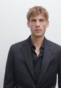 Schwarze Nadelstreifen-Sakko über einem schwarzen Hemd. Der Stoff wirkt glatt mit strukturierten Revers und einer Brusttasche. Einfaches, formelles Design.