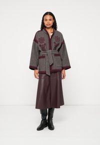 Γκρι μάλλινο σακάκι με μπορντό λουλουδάτη κεντητική, ζώνη στη μέση, φαρδιές μανίκια, συνδυασμένο με μπουρντό δερμάτινη φούστα και μαύρες μπότες μέχρι το γόνατο.