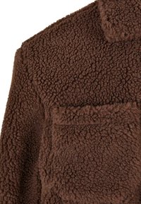 Brun fleecejakke med en struktureret overflade, krave og en frontlomme. Materialet er blødt og varmt med et shaggy udseende.