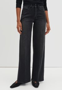 Personne portant un jean noir taille haute à jambes larges et des chaussures noires pointues avec des détails œillets, debout contre un fond uni.