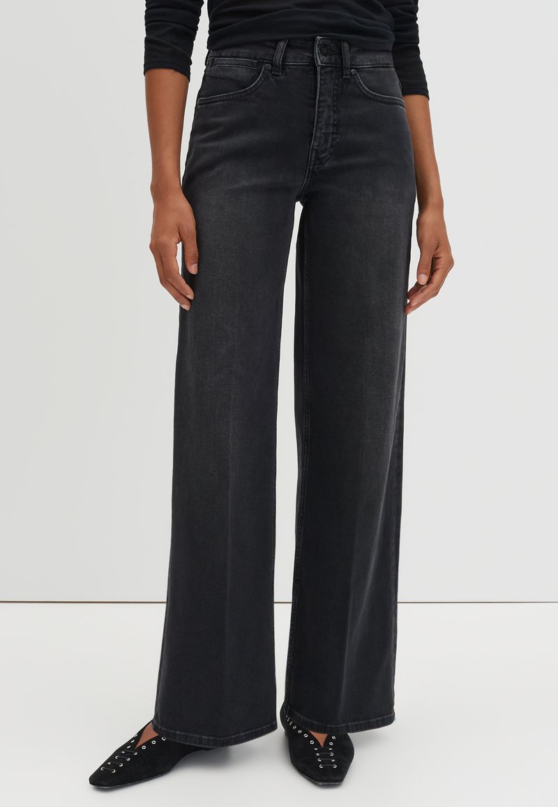 Personne portant un jean noir taille haute à jambes larges et des chaussures noires pointues avec des détails œillets, debout contre un fond uni.