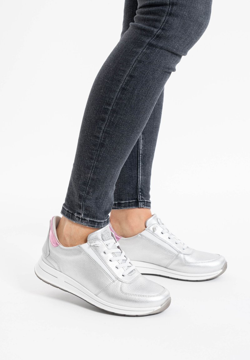 Zapatillas plateadas de cuero metálico, que presentan una suela blanca, diseño con cordones y acentos rosas en el talón, combinadas con jeans oscuros.