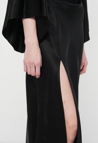 Melnas satīna kleitas ar zvana piedurknēm un augstu šķēlumu, izceļot gludo tekstūru un eleganto kritumu.