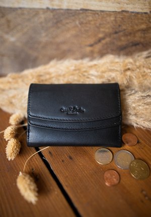 Musta nahkalompakko, jossa on teksturoitu pinta ja ommeltu yksityiskohta, varustettuna kohokuvioidulla logolla. Ympärillä kolikoita ja kuivattuja kukkia.