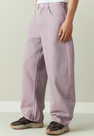 Person trägt locker sitzende lavendelfarbene Jeans und dunkelbraune Turnschuhe und steht auf einem hellen Boden vor einer schlichten Wand.