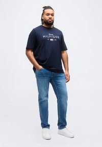 Άνδρας με κοτσίδες, φοράει μπλε μαρινιέρα Tommy Hilfiger, μπλε τζιν και λευκά αθλητικά παπούτσια, στέκεται με το ένα χέρι στην τσέπη.