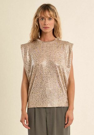 Donna con capelli biondi fino alle spalle che indossa un top beige senza maniche con stampa leopardata metallizzata e pantaloni grigi larghi, in piedi davanti a uno sfondo semplice.