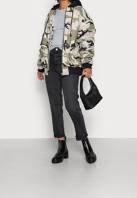 Kamelouttisate bomber-takki mustilla yksityiskohdilla harmaan toppatakin päällä, yhdistettynä tummiin farkkuihin ja mustiin nilkkureihin. Pieni musta käsilaukku kädessä.