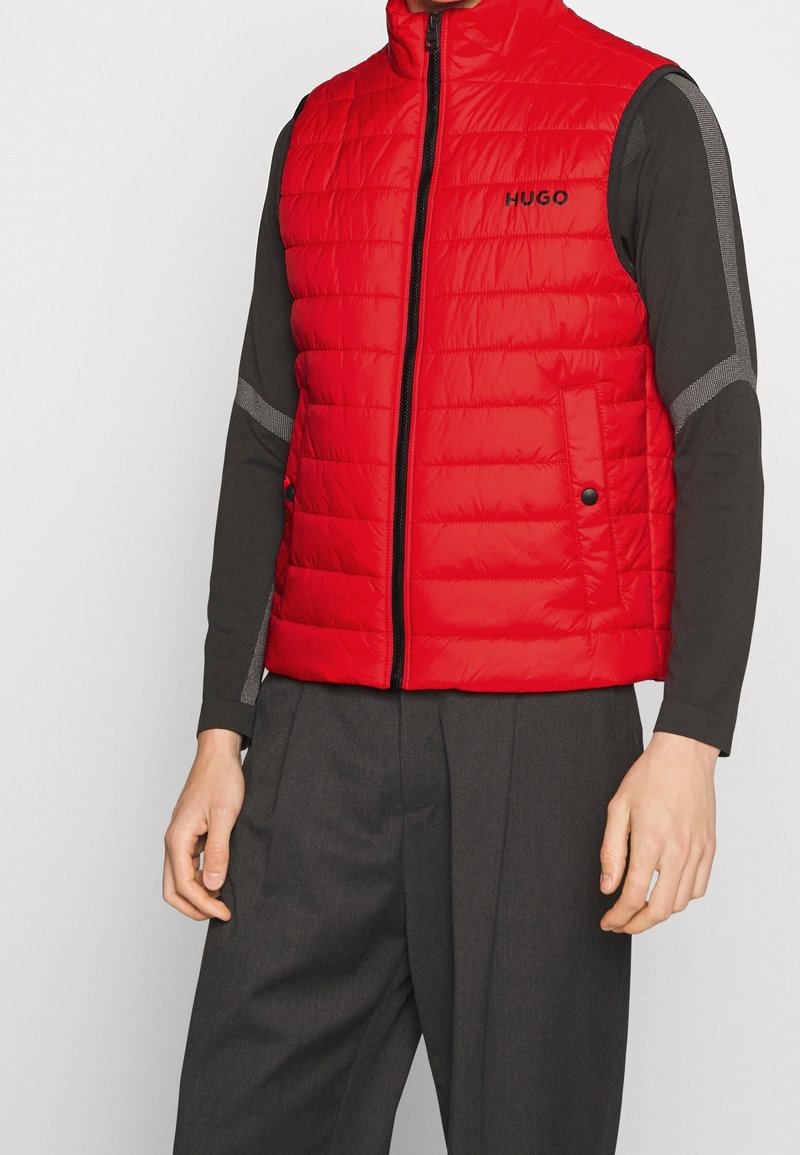 Rote gepolsterte Weste mit hohem Kragen, Frontreißverschluss und Logo. Verfügt über seitliche Taschen mit Knöpfen und wird mit einem dunklen Langarmshirt kombiniert.