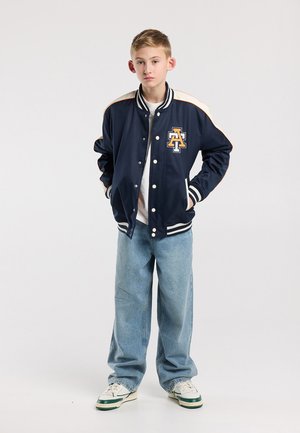 Garçon debout les mains dans les poches, portant une veste universitaire bleu et blanc, un jean bleu clair et des baskets blanches sur un fond uni.