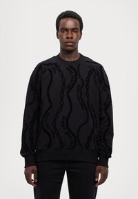 Felpa nera con stampa a rilievo tono su tono "Versace Jeans Couture" e motivo a catena. Presenta un collo a girocollo e polsini a coste, vestibilità relax.