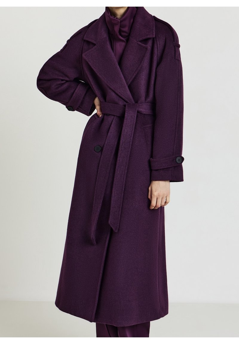Femme portant un long manteau en laine violet foncé ceinturé, avec des revers et des poignets boutonnés, debout avec une main sur la hanche.