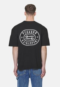 Fekete pamut póló, hátul grafikás logóval, kör alakú dizájnnal, rajta "PEGADOR" felirattal és kulcsokkal, rövid ujjú.