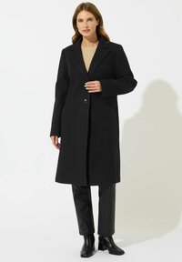 Cappotto di lana nero con un solo bottone, rever a pistagna e tasche laterali; indossato sopra un top beige e abbinato a pantaloni di pelle nera e scarpe.