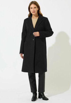 Abrigo de lana negro con un solo botón, solapas de muesca y bolsillos laterales; llevado sobre una blusa beige y combinado con pantalones de cuero negros y zapatos.