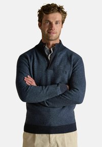 Tumši zila adīta džemperis ar ceturtdaļas rāvējslēdzēju un ribotu apkakli. Izceļas ar vidēju tekstūru un pieguļošu dizainu. Nēsāts ar svītrainu krekla apakšā.