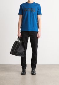 Camiseta de algodón azul con "BOSS" en negro, pantalones negros y un bolso tote negro texturizado con dos asas y cierre de cremallera. Zapatos negros.