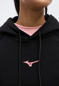 Κοντινό πλάνο προσώπου με μαύρη φούτερ με κουκούλα που φοράει με ροζ στυλιζαρισμένο λογότυπο και ροζ πουκάμισο από κάτω.