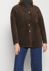 Chaqueta de pana marrón con un corte relajado, cierre de botones, dos bolsillos en el pecho y mangas largas, combinada con unos pantalones vaqueros de pernera ancha azules.