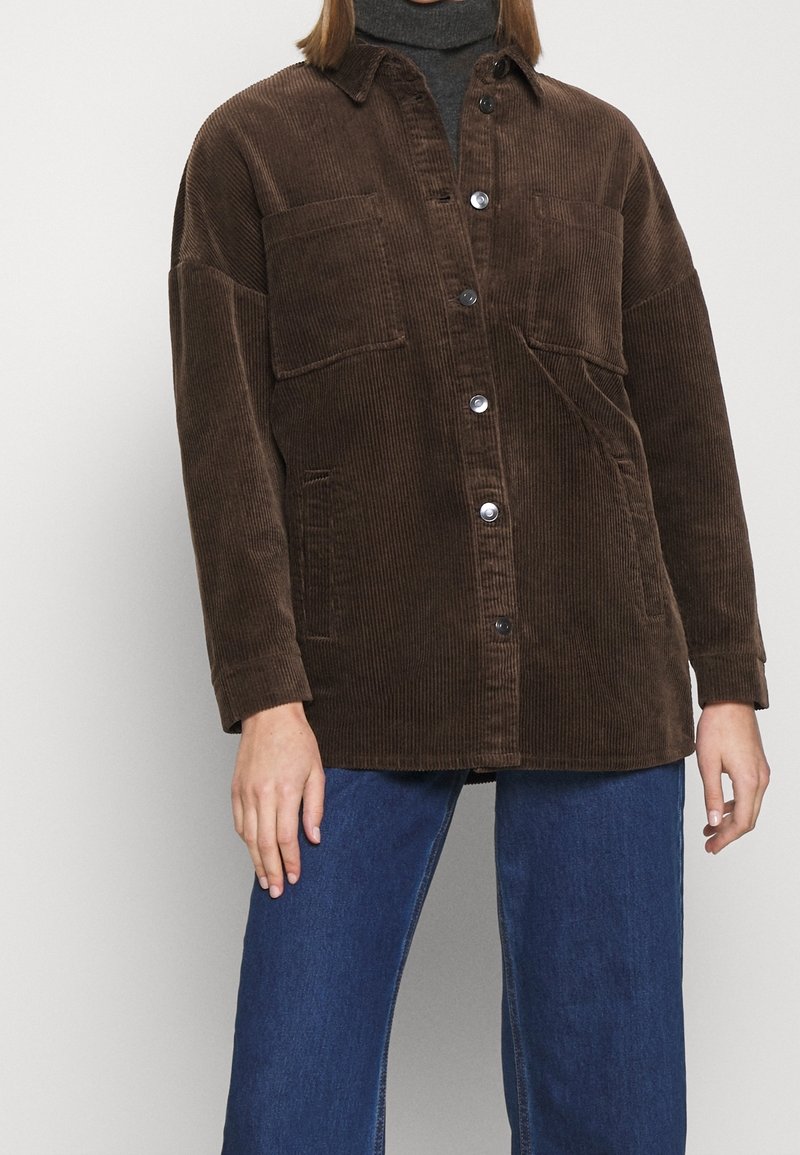 Chaqueta de pana marrón con un corte relajado, cierre de botones, dos bolsillos en el pecho y mangas largas, combinada con unos pantalones vaqueros de pernera ancha azules.