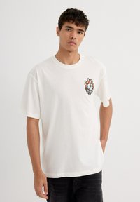 Camiseta de algodón blanca con un diseño gráfico que presenta llamas y un planeta en el lado izquierdo del pecho. Manga corta y corte relajado.