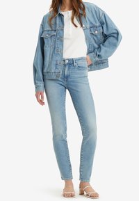 Světle modrá denimová bunda přes bílý žebrovaný top, spárovaná se světle modrými skinny džíny a bílými sandály na pásku, vystavená na neutrálním pozadí.