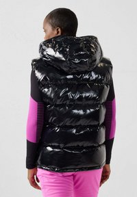 Smanicato imbottito nero lucido con cappuccio, presenta impunture orizzontali. Indossato sopra una maglia a maniche lunghe con accenti a righe rosa e maniche nere.