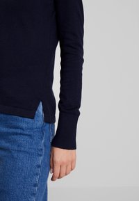 Navy long-sleeve sweater in soft material with ribbed cuffs. Features side slits at the hem and a fitted silhouette, paired with blue jeans.