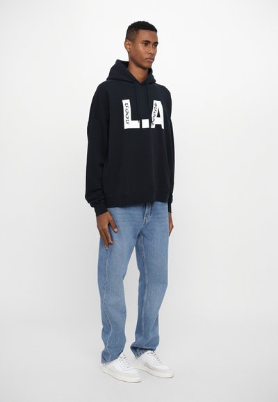 Schwarzer Hoodie mit weißem „L.A.“-Grafik, kombiniert mit hellblauen Jeans und weißen Sneakers. Aus Baumwollmaterial, lockerer Schnitt und minimalistisches Design.