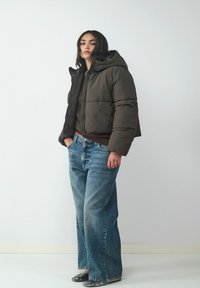 Gekürzte, dunkelgrüne Daunenjacke mit Kapuze, strukturierter Stoff, kombiniert mit weiten, hellblauen Jeans und schwarzen Schuhen.