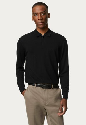 Camisa de polo negra de manga larga con cuello con cremallera, hecha de tela suave, combinada con pantalones beis y un cinturón negro.
