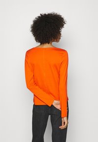 Orange langærmet top med rund halsudskæring og afslappet pasform, lavet af blødt stof. Kombineret med mørke, højtaljede jeans.