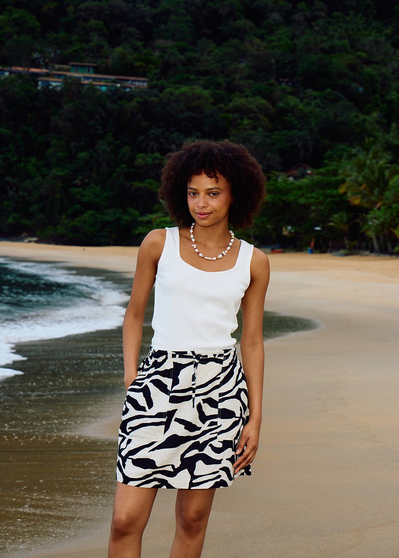 Jeune femme aux cheveux bouclés se tient sur une plage de sable, portant un haut blanc sans manches, une jupe à motif zèbre et un collier de perles.