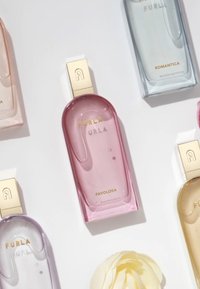 Fragranza FURLA Favolosa in una bottiglia di vetro rosa chiaro con un tappo dorato, insieme a bottiglie trasparenti e pastello, su uno sfondo bianco.