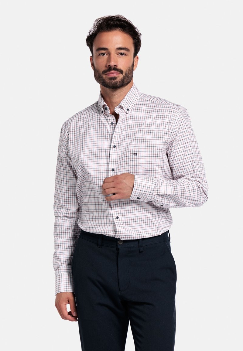 Man in een wit overhemd met een rood en blauw ruitpatroon en een donkere marineblauwe broek, poserend tegen een effen witte achtergrond.